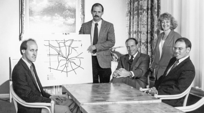 a black and white photo of a group of people in suits sitting around a table. one holds up a map/chart of a road system.