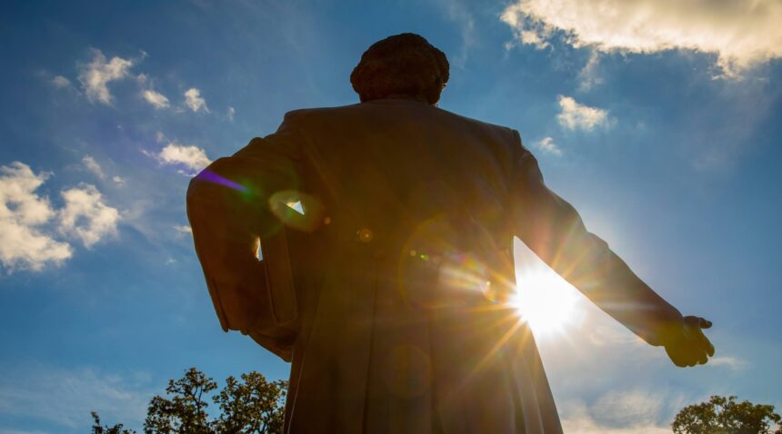 a photo of the statue of Matthew Gaines from behind with the sun in front, shooting beams of light under his outstretched arm