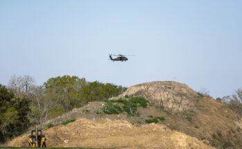 a photo of a large black helicopter flying over trees and a big pile of dirt