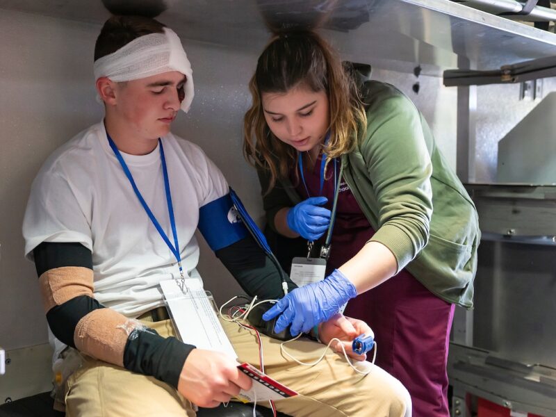 a photo of a young woman in maroon scrubs and a green jacket taking the pulse and blood pressure of another student dressed up in bandages and injury makeup