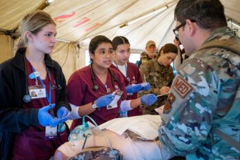 a group of female students in gloves and scrubs standing over a practice dummy with an oxygen mask on its face. The students are speaking with a man in a military uniform, while a woman in a military uniform writes on a clipboard in the background.