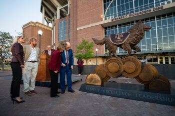 People stand to the side of a large bronze statue of Reveille in front of Kyle Field 