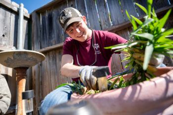 A male student wearing a baseball cap and maroon T-shirt kneels on the ground as he puts a plant in a flower pot