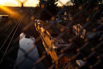 A train passes under a bridge at sunset, pictured behind a chain link fence in the foreground