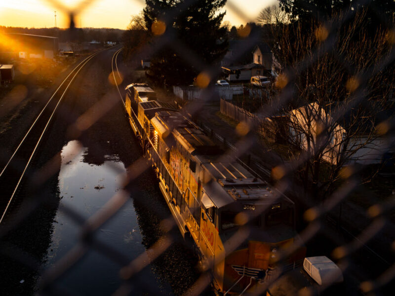 A train passes under a bridge at sunset, pictured behind a chain link fence in the foreground