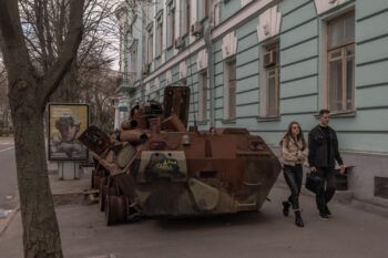 A photo of two young people, a man and a woman, walking past a light blue building facade and a rusted out armored vehicle with graffiti on it. In the background is an advertisement featuring a Ukrainian soldier in combat gear.