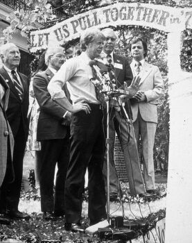 Black and white photo of Carter speaking in front of a microphone, with a banner stretching across the stage that reads "let us pull together"