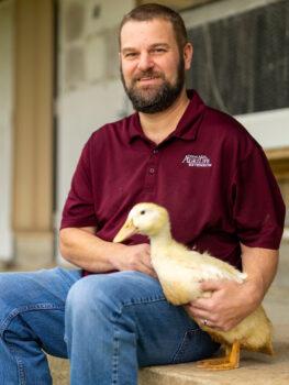 a photo of a bearded man in a maroon AgriLife polo and jeans sitting down and cradling an adult duck in his arm