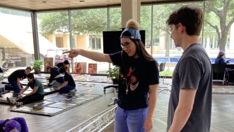 a photo of a young man and a young woman overseeing a group of students placing paintings and other works of art on boards for display