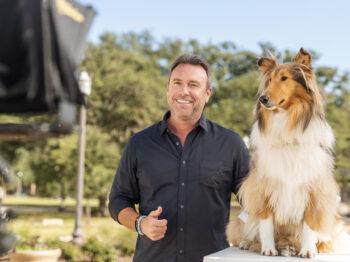 a photo of a middle-aged man in a black button-up shirt giving the thumbs up. he is standing next to a rough collie sitting on a pedestal.