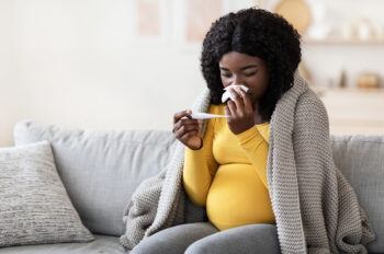 a pregnant woman wiping her nose with tissue and looking at a thermometer
