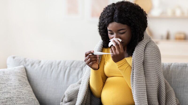 a pregnant woman wiping her nose with tissue and looking at a thermometer