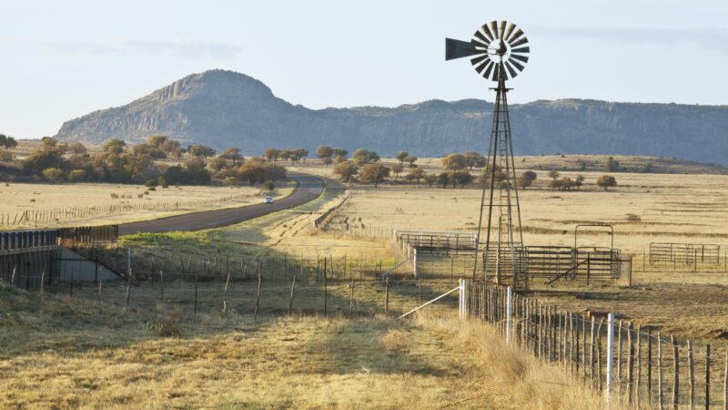 Fence-line leading to West Texas Ranch corrals and windmill, with highway running by. In early morning light.