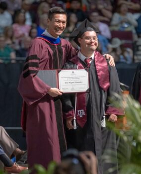A man in a maroon robe places his arm around a young man in a graduation gown and smiles, handing him a certificate