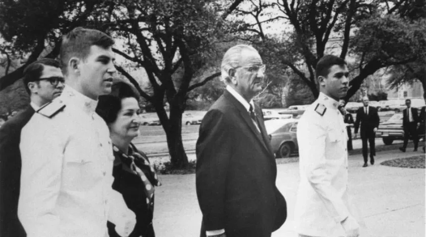a photo of Lady Bird and Lyndon Johnson accompanied by cadets in Ross Volunteer uniforms walking across the Texas A&M campus.