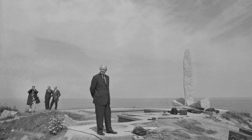 a photo of Dwight Eisenhower in a black suit standing near a stone monument on a cliffside, with various wires and recording equipment at his feet
