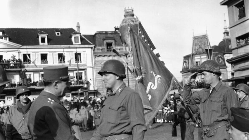 a black and white photo of a man in a french military uniform shaking hands with a man in an American military uniform in a town square as soldiers and civilians look on