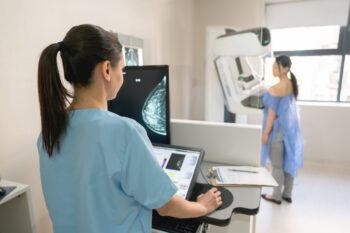 A nurse looking at a screen as a patient stands at a mammogram machine in an exam room
