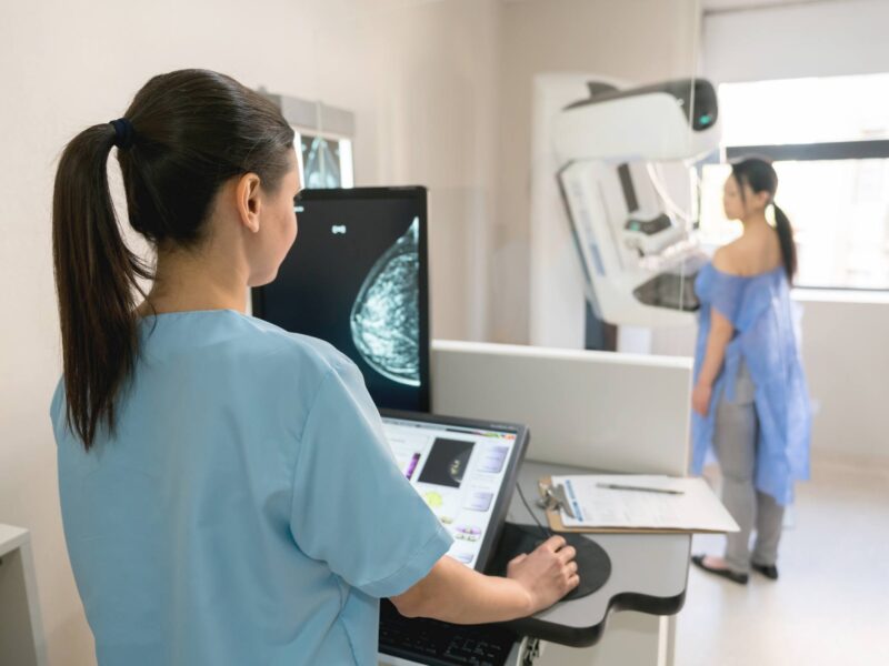 A nurse looking at a screen as a patient stands at a mammogram machine in an exam room