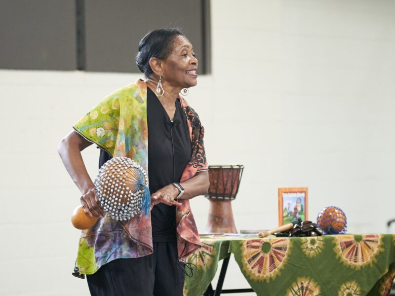 a photo of a woman holding a gourd instrument with a variety of instruments and other items behind her