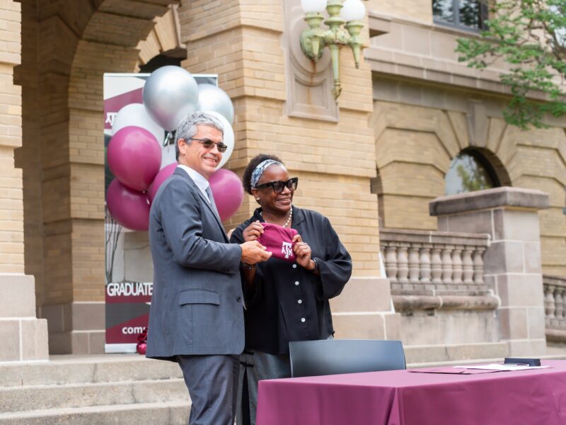 a photo of a woman in a black shirt holding an A&M shirt next to a man in a gray suit, with maroon and white balloons behind her