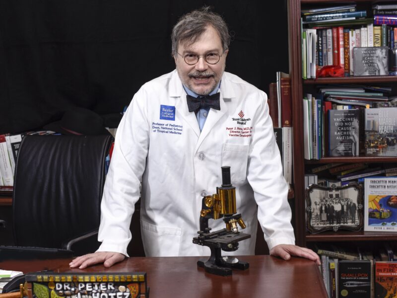 a portrait phot of a man with grey hair, glasses, a lab coat, and a bowtie standing at a desk with an old microscope in front of him