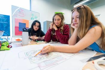 a photo of three young women looking at architectural plans on a table with a whiteboard and screen behind them