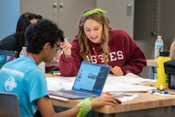 a photo of a young woman in an Aggies sweatshirt holding a pen and looking at plans on a table with several other young people