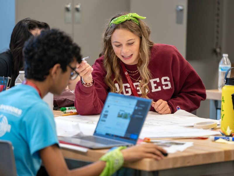 a photo of a young woman in an Aggies sweatshirt holding a pen and looking at plans on a table with several other young people