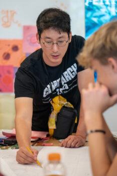 a photo of a man with round glasses and a Camp ARCH t shirt pointing at architectural plans on a table while a student in the foreground looks on