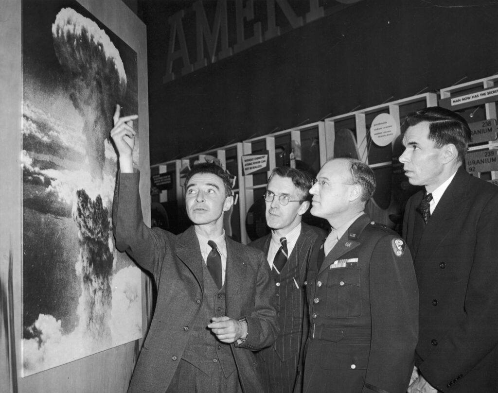 a black and white photo of a group of men looking at a picture of a mushroom cloud, as the man next to the picture gestures towards the top of the cloud