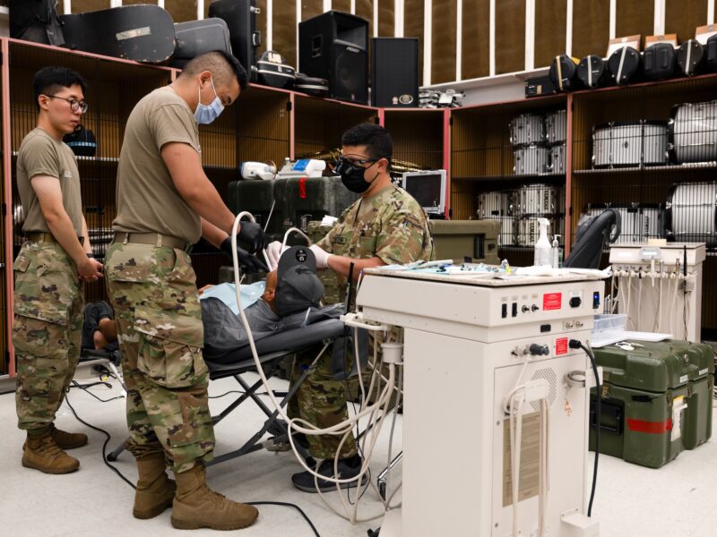 A photo of several people in army fatigues using various dental instruments a reclining man. In the background, drums and other instruments can be seen inside lockers.