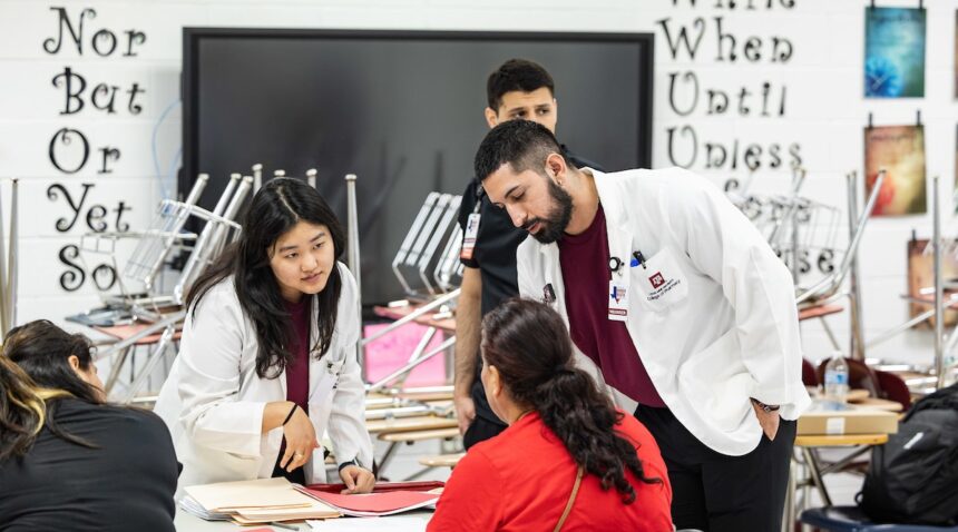 a photo of several students in maroon shirts and lab coats standing around a table talking to a woman in a red shirt
