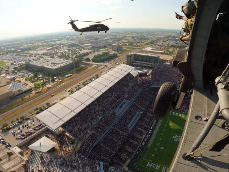 a still from a video showing a man in a flight helmet throws up a gig 'em while flying over Kyle Field. The stands below are packed with fans wearing maroon and white, and a second helicopter can be seen in the near distance