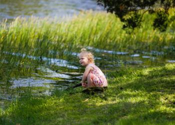 A photo of a young girl kneeling in the grass next to a lake.