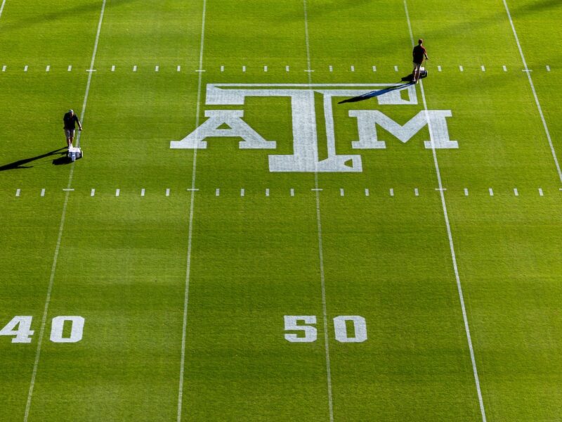 overhead view of kyle field. A man on the 50 yard line paints the A&M logo in white paint onto the turf grass.