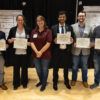 A photo of six people. Five of the people are holding awards received during a campus symposium.