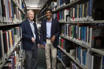 A photo of two men standing among shelves of books in a library.