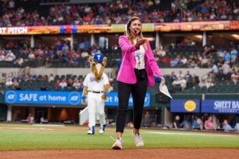 A woman in a pink blazer walks across a baseball field holding out her hand. The mascot of the Texas Rangers, Ranger Captain (a large fuzzy horse in a Rangers uniform), can be seen behind her