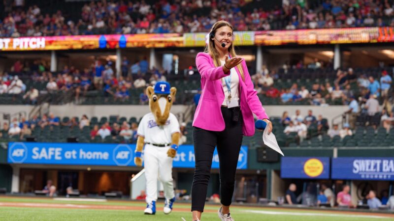 A woman in a pink blazer walks across a baseball field holding out her hand. The mascot of the Texas Rangers, Ranger Captain (a large fuzzy horse in a Rangers uniform), can be seen behind her