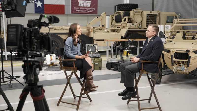 Host Chelsea Reber seated in a chair interviewing Tim Green with military vehicles set up in the background