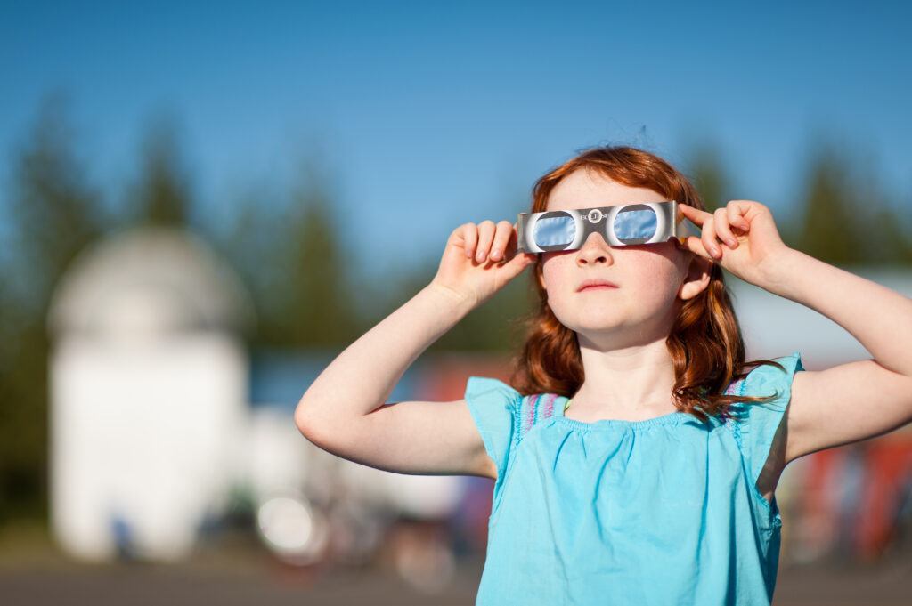 A young girl with red hair watches a solar eclipse with special viewing glasses.