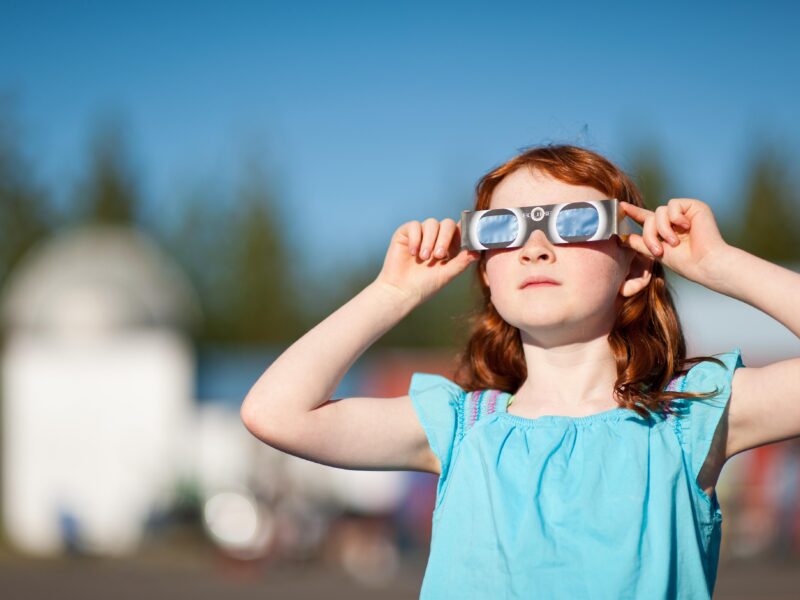 A young girl with red hair watches a solar eclipse with special viewing glasses.