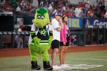 Dallas Stars mascot Victor E. Green (a large green fuzzy monster wearing a Rangers jersey on top of a his Stars jersey) stands next to a man in a rangers jersey and a woman in a pink blazer