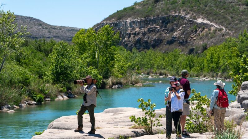 A group of five people take photos and enjoy the scenery along a river in a semi-arid Texas landscape.