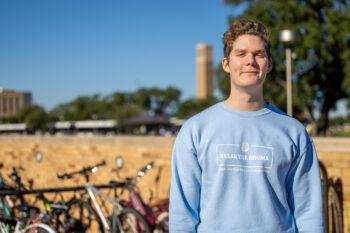 a young man in a blue sweatshirt that reads "break the stigma" stands in front of a bike rack with the Albritton Bell Tower in the background.