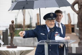 Buzz Aldrin standing at a lectern in a U.S. Air Force uniform giving remarks while a man stands behind him holding an umbrella over him.