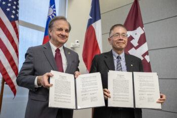 a photo of two men holding up signed papers in front of the U.S. flag, the Taiwan flag, the Texas flag and the Texas A&M flag.