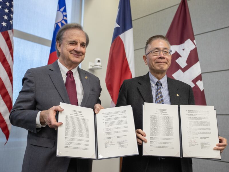 a photo of two men holding up signed papers in front of the U.S. flag, the Taiwan flag, the Texas flag and the Texas A&M flag.