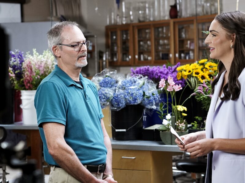 a photo of a man and woman talking with a table filled with colorful flowers behind them